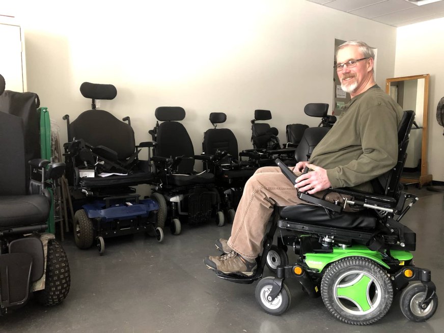 An older white man in an olive green shirt and khaki pants sitting in a motorized wheelchair, smiling for the camera. Behind him is a row of other motorized wheelchairs.