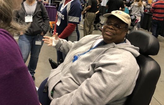 An African American woman wearing a white hoodie and baseball cap gestures as she talks to an unseen group, while sitting in a mobility device.